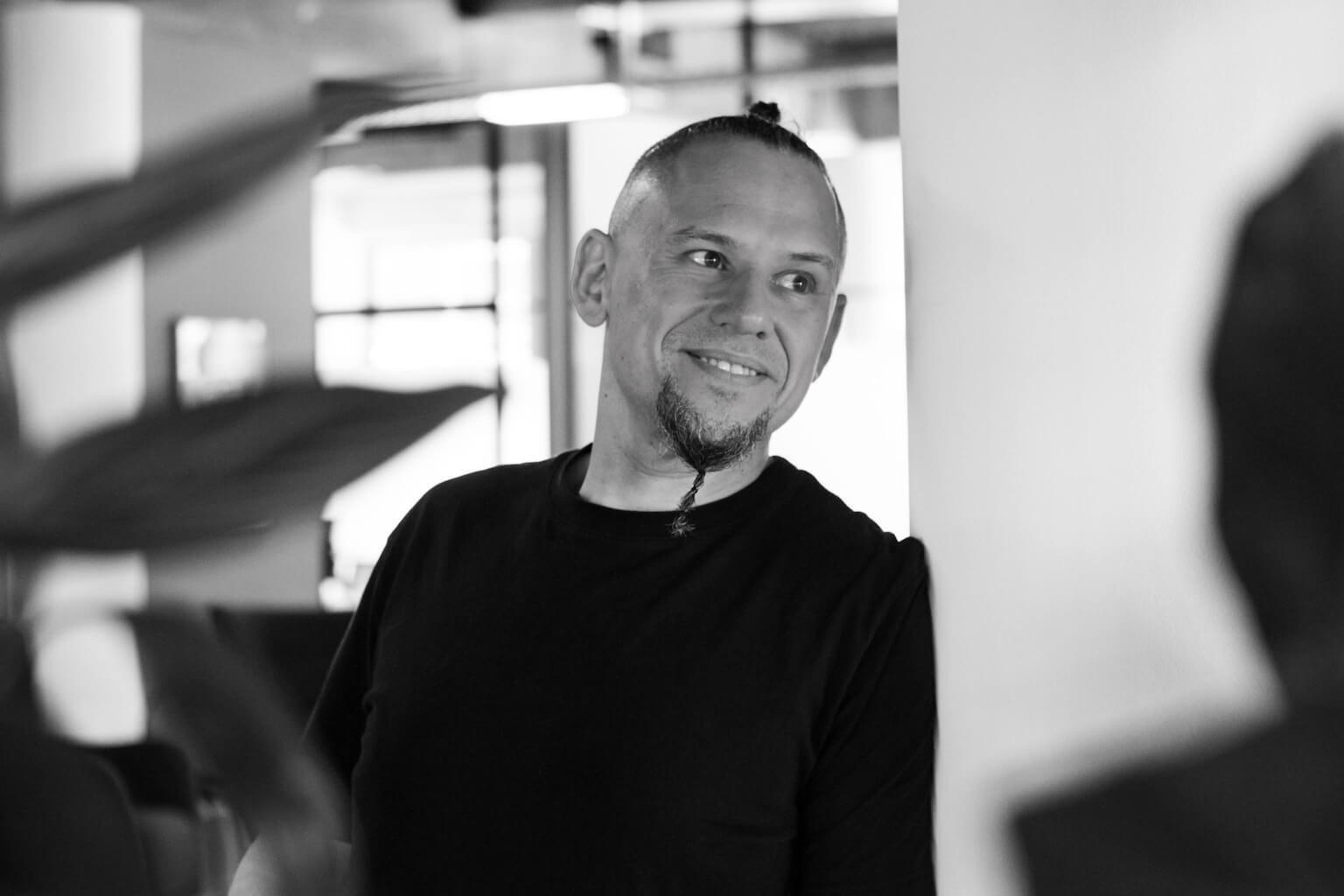 Andreas Schmitz Blick zur Seite Die Person im Bild, Andreas Schmitz, blickt angelehnt an eine Wand zu Seite, trägt ein schwarzes T-Shirt und lächelt.